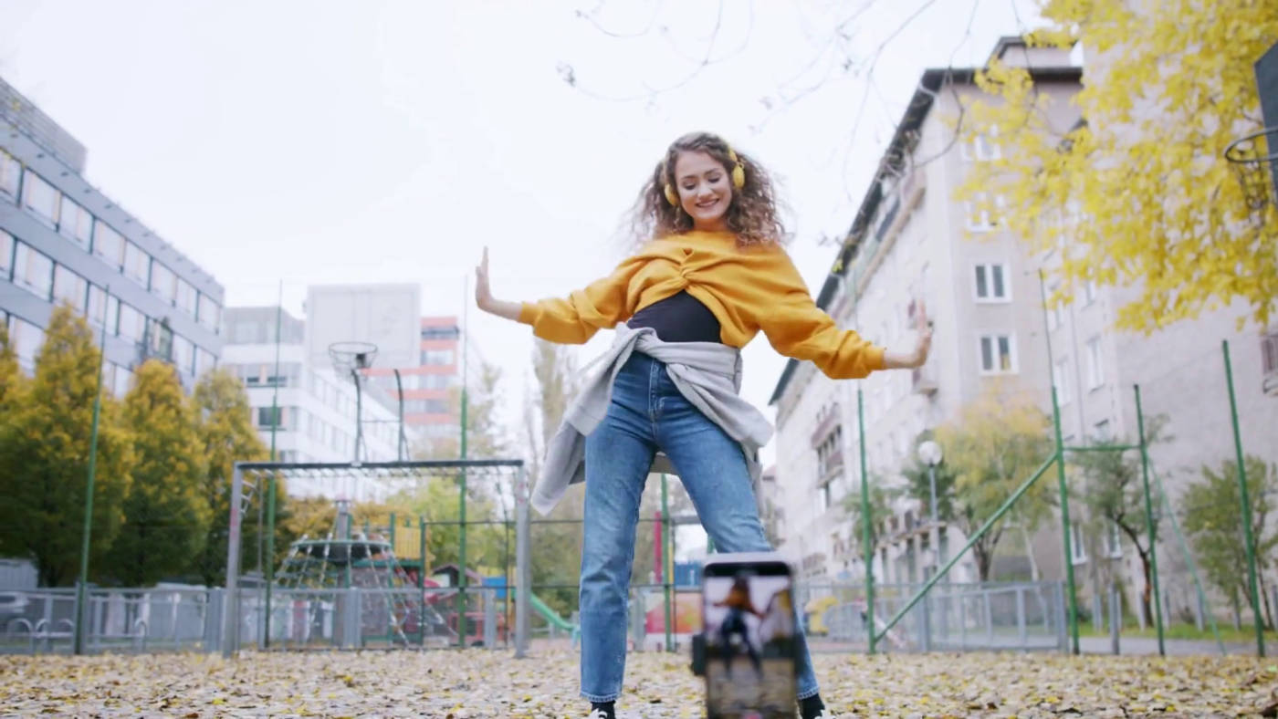 A woman dressed in yellow top and jeans dancing outside in front of a smartphone