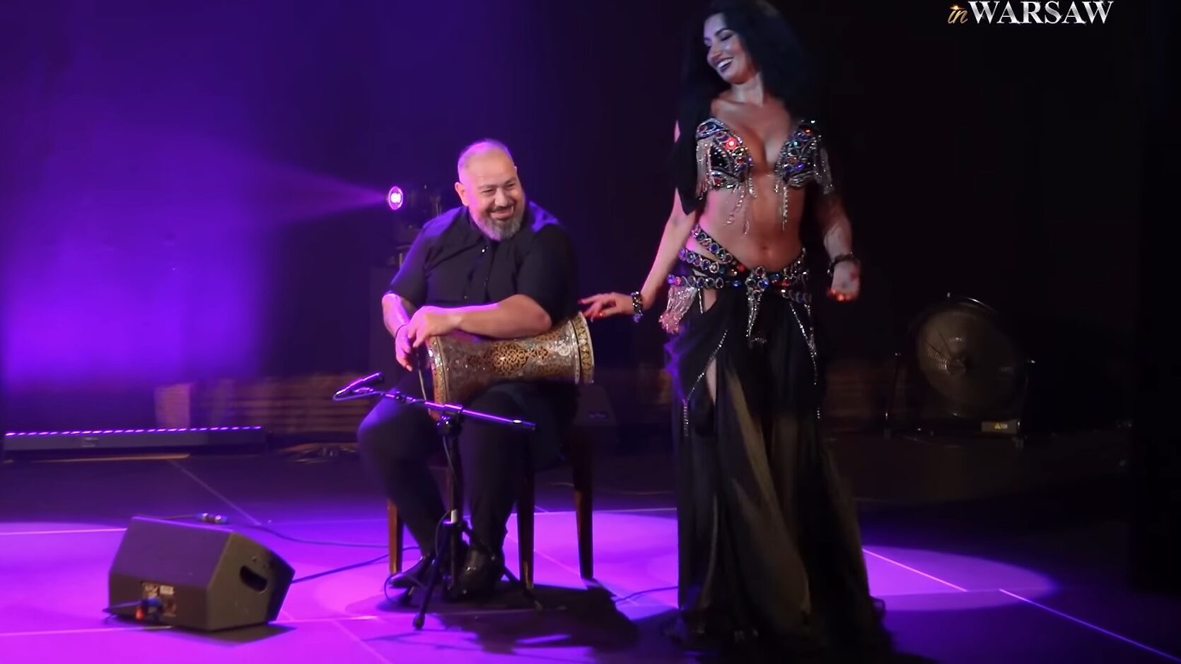A belly dancer woman and a drum solo musician on stage smiling at each other
