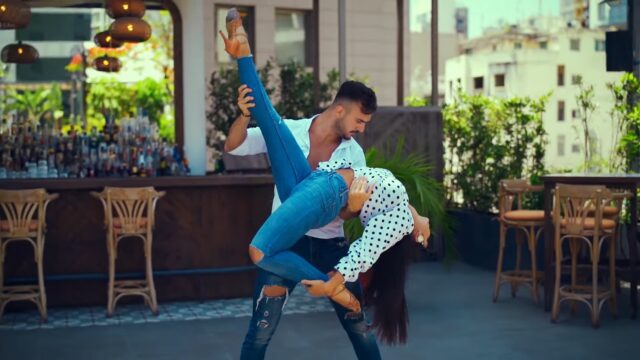 Man in white shirt and blue jeans holding woman a polka dot blouse and blue jeans in an acrobatic lift after dancing salsa near a cafe