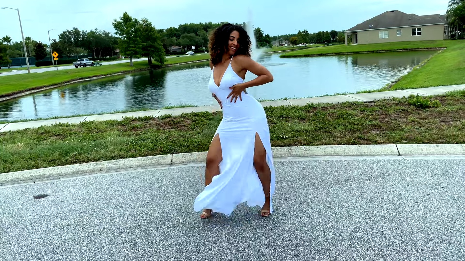 A woman in white dress dancing bachata outisde near a pond with green lawn
