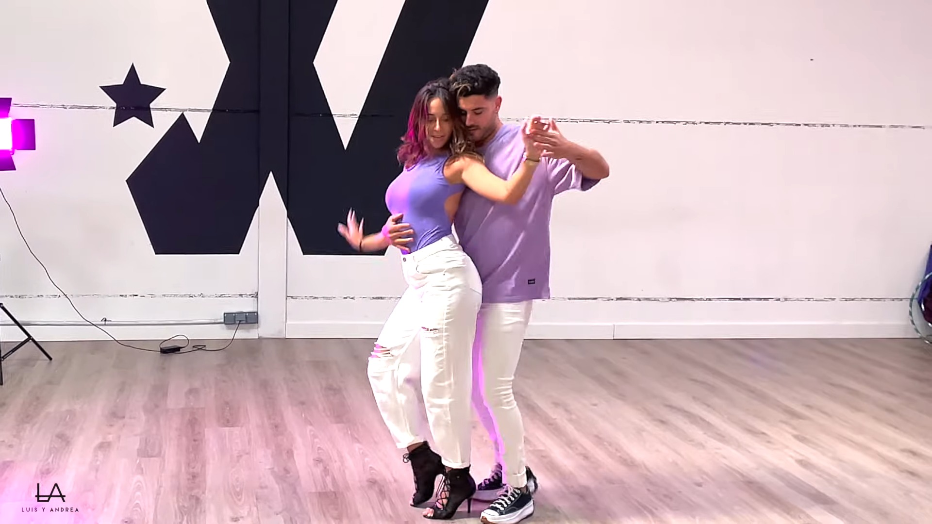 A man and a woman in white pants and purple tops dancing together bachata in a class.