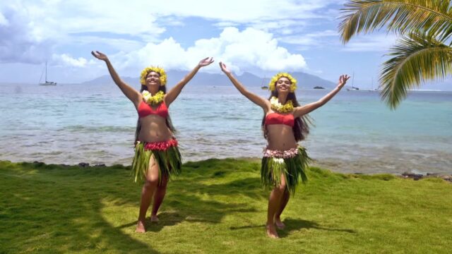 Two women in traditional Tahitian costumes dancing near the ocean with the beautiful tropic scenery in the background