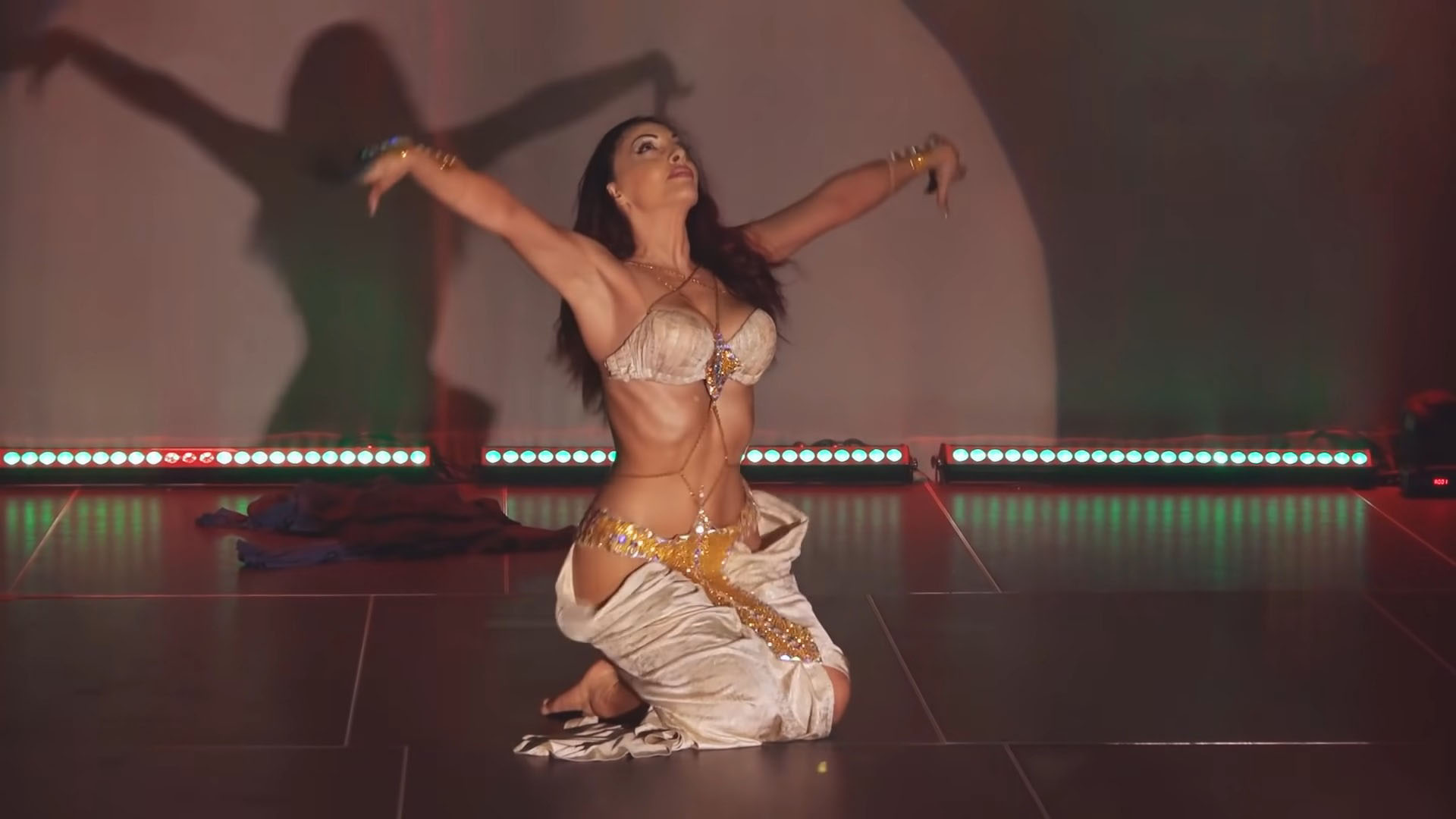 Woman sitting on stage with her arms raised looking up while belly dancing