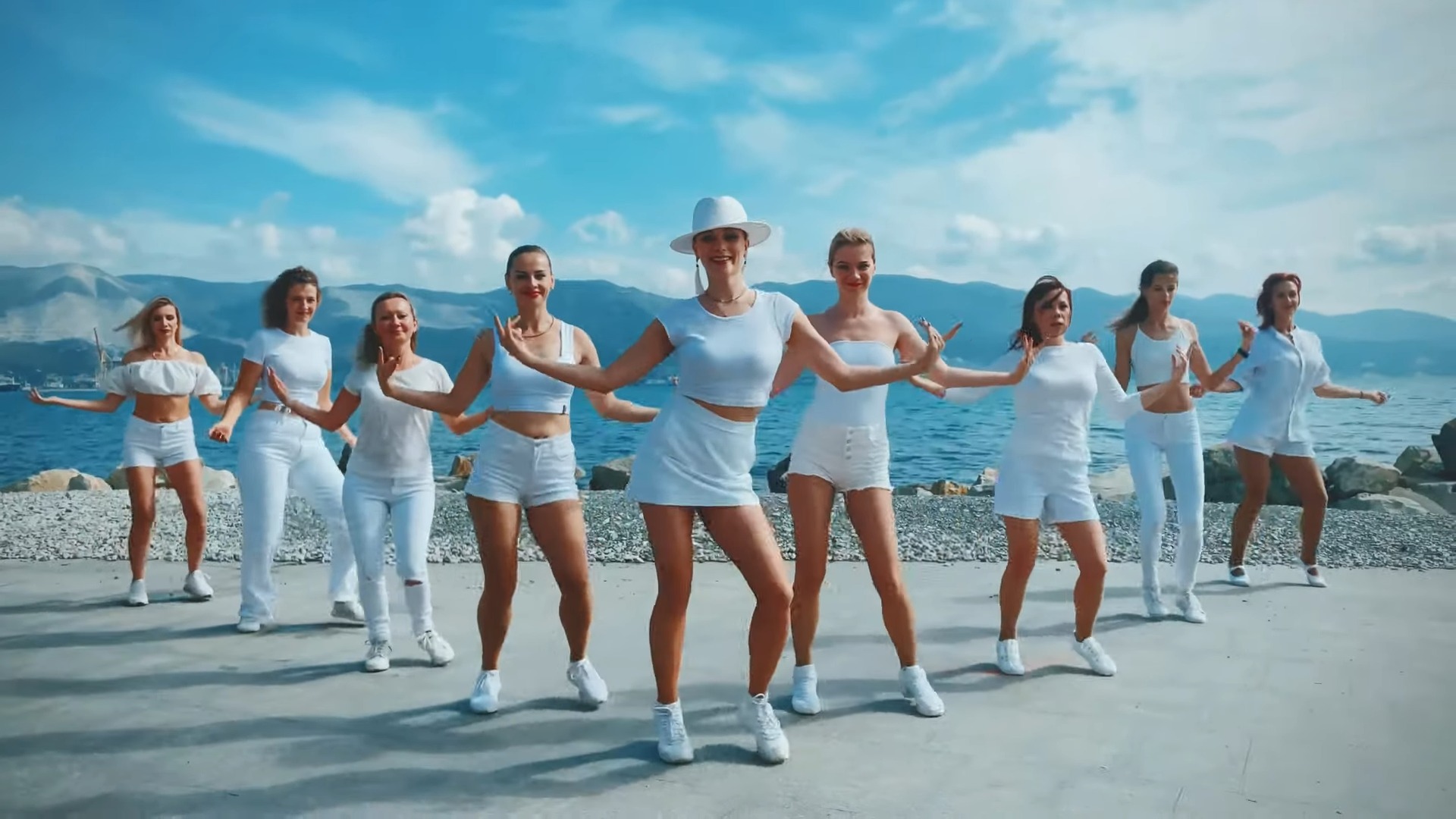 Women in white smiling and dancing bachata near the sea and mountains in the background